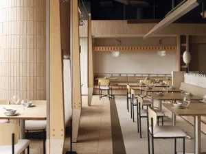 Modern minimalist Japanese restaurant interior featuring light oak wood paneling, concrete accents, rows of wooden tables with beige upholstered chairs, and layered pendant lights.