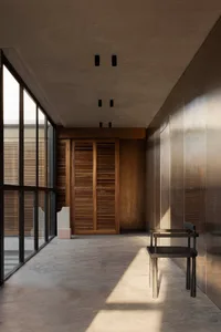 Minimalist concrete hallway featuring a dark walnut louvered cabinet, black wooden chair, large glass windows, and warm sunlight creating strong diagonal shadows.