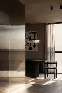 Modern minimalist dining room featuring a large brushed stainless steel room divider, dark wood dining table, woven chair, abstract painting, and a long cylindrical pendant light with natural sunlight streaming in.