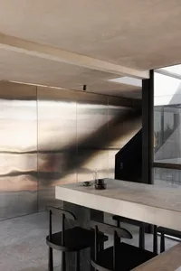 Minimalist dining area featuring a raw concrete island, dark wooden stools, and a large rippled stainless steel feature wall under a concrete ceiling.