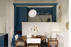 Modern dining area featuring a round walnut table, white canvas dining chairs, bold blue built-in cabinetry, and a marble kitchen backsplash in the background.