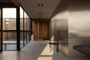 Modern minimalist hallway featuring polished concrete floors, large black-framed windows, wooden louver doors, a brushed stainless steel wall, and a dark metal chair bathed in natural sunlight.
