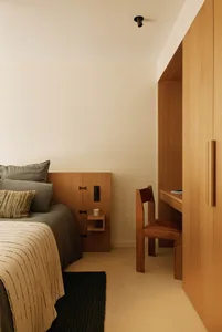 Modern minimalist bedroom featuring light oak built-in headboard, floating nightstand, integrated desk and chair, with neutral linen bedding and dark textured rug.