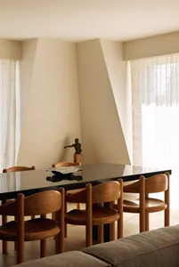 Modern minimalist dining room featuring six curved walnut wooden chairs, a long black reflective dining table, a bronze sculpture on a wooden pedestal in the corner, and soft sheer curtains allowing diffused daylight.