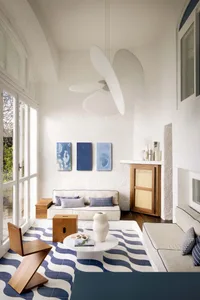 Bright sunroom interior with blue and white wave-pattern terrazzo floor tiles, white sofa, wooden zigzag chair, round marble coffee table, and large white sculptural pendant lamp.