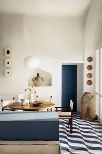 Bright dining room featuring wavy blue and white terracotta floor tiles, round wooden dining table, wooden chairs with striped cushions, a large white pendant lamp, and a blue door in the background.