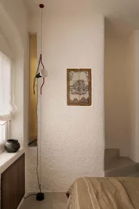 Minimal bedroom corner featuring heavily textured white plaster walls, a red and black adjustable pendant lamp, an antique four-tile decorative panel with botanical illustration, a dark wood cabinet, and a bed with neutral linen bedding.