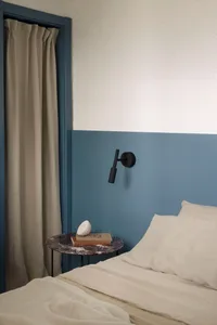 Modern bedroom corner featuring a two-tone blue wall, black wall sconce, round purple marble side table with books and a lamp, and a bed with neutral linen bedding.