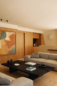 Modern minimalist living room featuring light oak wood paneling, a large abstract painting, a low dark charred wood coffee table, a neutral fabric sofa, and a woven jute rug under warm ambient lighting.