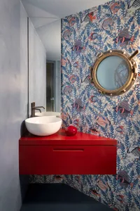 Modern powder room with vibrant fish and coral wallpaper, brass porthole mirror, red floating vanity, and white vessel sinks