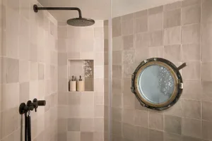 Modern bathroom shower corner with large round brass porthole mirror, beige rectangular tiles, black rain shower head and matte black wall mixer.