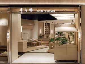 名為「斗山睡 under/clouds」的現代日式餐廳入口，採用大量淺木色板材、嵌入式座位、豐富綠植與層次柔和燈具。