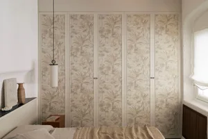 Modern bedroom interior showing a bed with neutral linen bedding, a wooden nightstand, and a large wardrobe covered in beige and taupe tropical leaf patterned textile wallpaper, with a cylindrical pendant lamp hanging above.