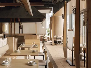 Modern minimalist restaurant interior featuring light plywood tables and chairs, upholstered banquettes, raw concrete ceiling elements, and large windows allowing abundant natural light.
