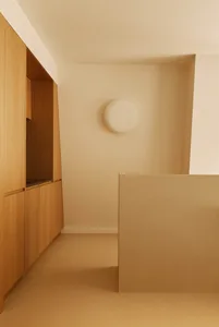 Minimalist entryway featuring light oak built-in cabinetry, hidden storage, a round white wall sconce, and a beige half-wall partition.