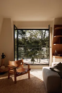 A contemporary living room featuring a wooden and tan leather armchair on a woven jute rug, opening to a balcony with green metal patio furniture overlooking a dense forest.