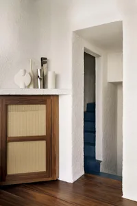 Modern minimalist entryway featuring a dark walnut cane cabinet displaying ceramic and metallic decor, adjacent to a textured white wall opening revealing a bold navy blue staircase.