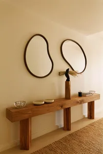 Minimalist entryway featuring a thick reclaimed wood console table, two large irregular organic-shaped mirrors with dark wood frames, and a sculptural bronze figure on a wooden pedestal.