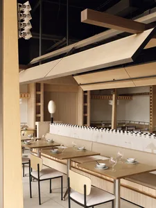 Modern minimalist restaurant interior featuring light oak wooden tables, upholstered booths, black metal chairs with bentwood backs, and a dramatic sculptural oak canopy hanging from the ceiling.