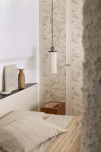 Cozy bedroom corner with beige linen bedding, wooden nightstand, cylindrical pendant lamp, and tropical leaf wallpaper in neutral tones.