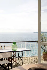 Outdoor stone balcony with white folding chairs, green glass vase, and sweeping ocean view under bright daylight