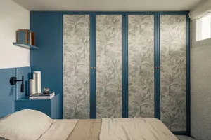 Modern bedroom with deep blue walls, a built-in wardrobe covered in grey tropical leaf wallpaper, a floating blue shelf with books, and a bed with neutral linen bedding.