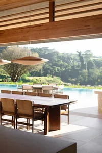Modern open-air dining area with long teak table and upholstered chairs, overlooking an infinity pool and lush green hills under a wooden ceiling with pendant lights.