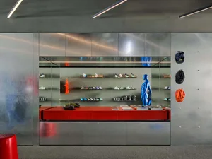 Modern sneaker retail display wall with stainless steel panels, red bench, floating shelves of sneakers, blue male mannequin sculpture, and baseball caps hanging on the right wall.