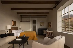 Modern mountain lodge bedroom featuring exposed dark wood ceiling beams, a bed with mustard yellow throw, beige sofa, dark coffee table, and large window overlooking snowy mountain landscape.