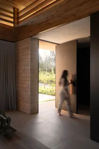 Modern minimalist entryway featuring a textured limestone wall, light wood flooring, warm wooden ceiling with integrated lighting, and a large pivot door opening to a garden, with a person walking through.