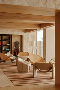 Modern living room with beige boucle sofas, rattan lounge chairs, a striped area rug, stone walls, and wooden ceiling beams.