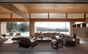 Modern open-plan living room featuring a massive wooden slat ceiling, large sectional sofa, mid-century lounge chairs, and expansive glass walls with pool and mountain views.