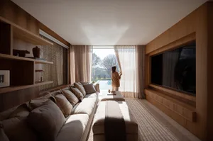 Modern living room featuring extensive walnut wood cabinetry and built-in TV wall, a large neutral sectional sofa, and a woman opening sheer curtains to a sunlit pool and garden view.