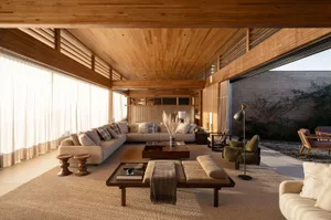 Spacious modern living room featuring extensive wood paneling on ceiling and walls, a large L-shaped beige sectional sofa, wooden coffee tables, a green swivel armchair, and direct view to an outdoor courtyard with brick wall.