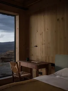 Cozy mountain cabin bedroom featuring light wood panel walls, a dark wood writing desk, woven rattan chair, and large window overlooking a dramatic landscape.