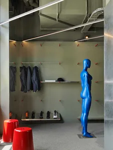 Modern athletic locker room with sage green walls, stainless steel benches and hooks, hanging dark sportswear, a row of sneakers, two glossy red stools, and a full-body glossy blue mannequin standing on the right.