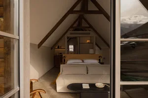 Cozy attic bedroom with exposed dark wood ceiling beams, neutral linen bedding, black coffee table, and large windows overlooking snowy mountain landscape.