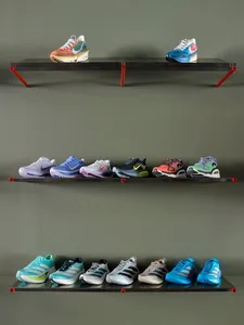 Three rows of colorful running sneakers from Nike, Adidas, and On displayed on black floating shelves with bright red metal brackets against a muted green wall.