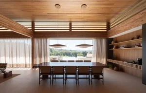 Modern minimalist dining room featuring extensive oak wood ceilings and walls, a long dark dining table with eight upholstered chairs, two large elliptical pendant lights, and large windows revealing a mountain and pool view.
