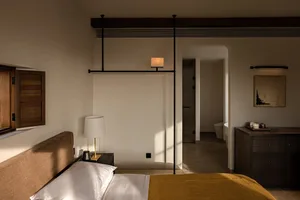 Modern minimalist bedroom with brown upholstered bed, mustard yellow throw, walnut furniture, and black sliding door leading to an ensuite bathroom