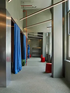 Modern industrial-style retail changing room featuring bright blue curtains, exposed red pipes, concrete walls, grey carpet, and colorful plastic stools with a blue mannequin.