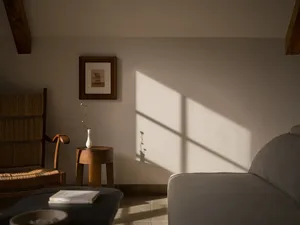 A tranquil living room corner featuring a wooden rocking armchair with woven seat, a round wooden side table with a white vase, and soft morning sunlight creating strong diagonal shadows on a neutral wall.