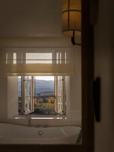 White freestanding bathtub in front of a large open window with oak frame, overlooking autumn mountains and vineyards, with a woven pendant lamp hanging nearby.