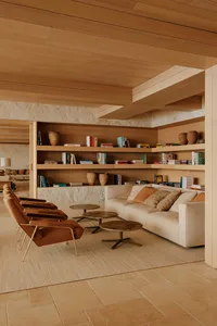 Modern living room featuring built-in wooden bookshelves, a travertine stone wall, a cream sofa with textured pillows, four terracotta leather lounge chairs with brass legs, and round wooden coffee tables on a large area rug.