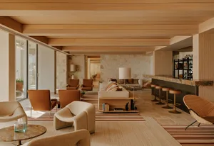 Spacious modern Mediterranean living room featuring travertine stone walls, oak wood ceiling, terracotta leather armchairs, cream bouclé sofas, and a marble-topped bar with rattan bar stools.