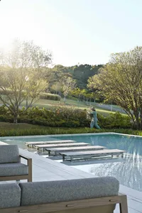 Modern infinity pool with teak and fabric lounge chairs on a stone deck, a person tending to plants in the background with forested hills beyond.