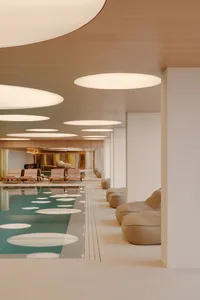Modern indoor swimming pool with teal water, wooden ceiling, large circular pendant lights, and beige lounge chairs and beanbags.