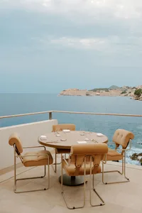 Round outdoor dining table with four mustard-ochre upholstered chairs on a stone balcony overlooking the Adriatic Sea and the old town of Dubrovnik, Croatia.
