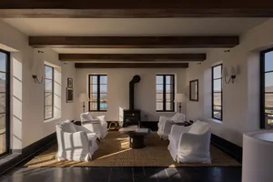 Bright modern farmhouse living room featuring white slipcovered armchairs, a round black coffee table, exposed wooden ceiling beams, black steel windows, and a wood-burning stove with mountain views.