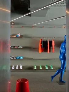 Modern sneaker retail display wall with colorful athletic shoes on clear acrylic shelves, bright orange and black apparel hanging on a clothing rack, and a glossy blue mannequin standing in the foreground.